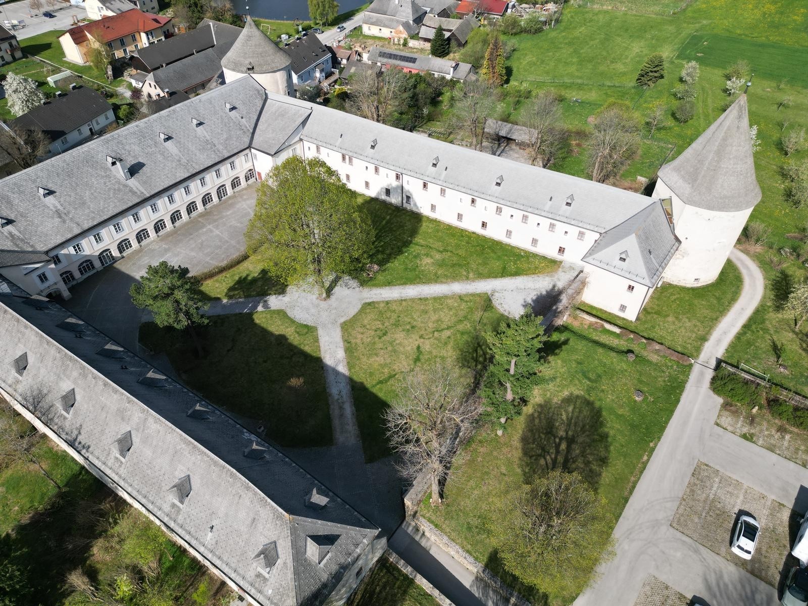 Ein Bild vom Schloss von oben. Es ist zu sehen, dass es ein großer offener Dreikanthof ist. An drei Ecken befinden sich Türme. Im Hof sieht man mehrere Schotterwege sowie einen großen Baum im Zentrum.